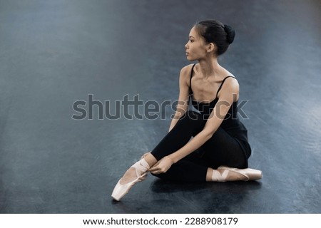 An Asian woman dressed in a black bodysuit, leggings and pointe shoes sits on the floor.