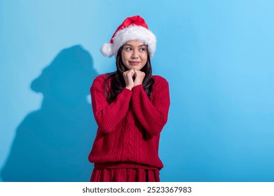 An Asian woman dressed in a red sweater and Santa hat looks playfully to the side with her hands near her chin. The background is light blue, creating a festive and fun holiday atmosphere - Powered by Shutterstock