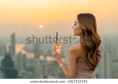 Asian woman drinking cocktail with looking cityscape at skyscraper rooftop bar in the city at summer sunset. Attractive girl enjoy urban outdoor lifestyle hanging out nightlife on holiday vacation.