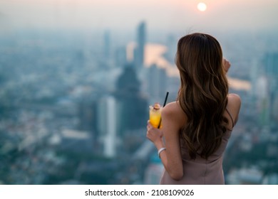 Asian Woman Drinking Cocktail While Waiting For Meeting With Friends At Skyscraper Rooftop Restaurant In Metropolis At Summer Sunset. Beautiful Female Enjoy Outdoor Lifestyle In The City At Night