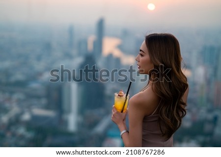 Asian woman drinking cocktail while waiting for meeting with friends at skyscraper rooftop restaurant in metropolis at summer sunset. Beautiful female enjoy outdoor lifestyle in the city at night