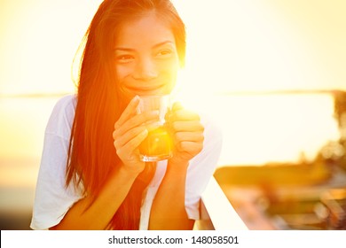 Asian woman drinking coffee in sun sitting outdoor in sunshine light enjoying her morning coffee. Smiling happy multiracial female Asian Chinese / Caucasian model in her 20s.