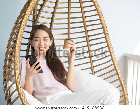 Asian woman drinking coffee and using smartphone while sitting in modern armchair in shape of birds nest at home