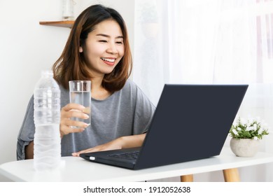 Asian Woman Drinking Fresh Water At Work Place Sitting At Office Hand Typing On Laptop And Holding Glass Of Water 