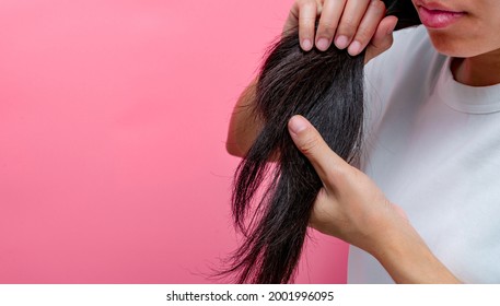 Asian woman with dry lips holding damaged hair on pink background. Dry and brittle black long hair needs shampoo and conditioner for spa treatment. Hair loss and thin hair problems in women. 