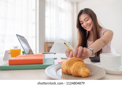 Asian Woman Eating Croissant While Working With Laptop On Table At Home.work At Home Concept