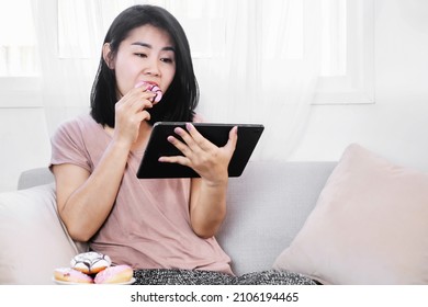 Asian woman eating donut and watching series on tablet sitting on sofa , unhealthy lifestyle and sugar addict concept 