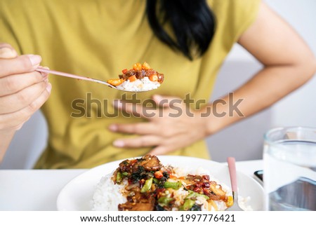 Asian woman eating spicy food and having acid reflux or heartburn hand holding a spoon with chili peppers another hand holding her stomach ache 