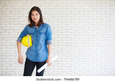 Asian woman engineer in blue jeans shirt carrying paper roll standing in front of brick wall, holding yellow safty helmet in self confident manner