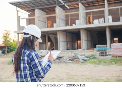 Asian Woman Engineer Fill Out Form Or Inspect In Front Of Construction Site. Civil Engineer Pointing, Writing, And Checklist At Building Project. Engineer And Worker Inspect Concept.