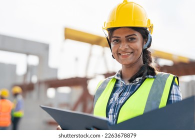 Asian woman engineer holding document smiling at construction site. Confident female Indian wearing protective helmet and vest working in factory making precast concrete wall for real estate housing. - Powered by Shutterstock