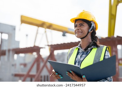 Asian woman engineer holding document smiling at construction site. Confident female Indian wearing protective helmet and vest working in factory making precast concrete wall for real estate housing. - Powered by Shutterstock