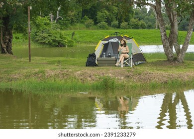 An Asian woman is enjoying sitting near a lake and drinking hot coffee in nature while in a tent camp outdoors camping. - Powered by Shutterstock