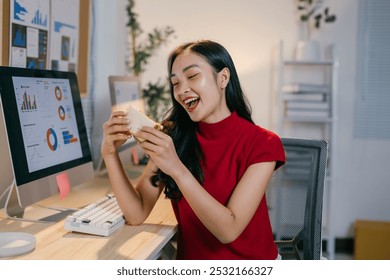 Asian woman enjoys a healthy lunch at her desk in a modern office, smiling happily as she eats a sandwich and uses her computer, taking a break from work - Powered by Shutterstock