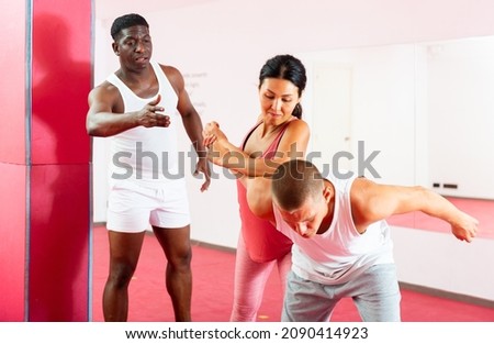 Asian woman exercising joint lock move together with caucasian man in gym during self-defence training. African-american man as a trainer standing beside and correcting them.