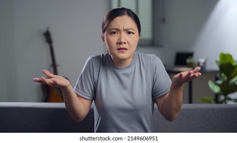 Asian Woman Feel Confused Looking At Camera Shrugging And Sitting In Living Room At Home.