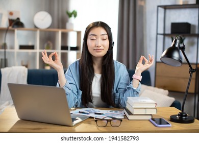 Asian woman freelancer in headset sitting at table with closed eyes and relieving stress by meditation at workplace. Concept of relaxation and harmony, no stress free relief at work