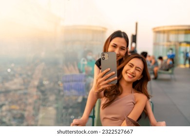 Asian woman friends enjoy urban lifestyle using mobile phone taking selfie together during have celebration dinner party at skyscraper rooftop bar in the city at sunset on summer holiday vacation. - Powered by Shutterstock