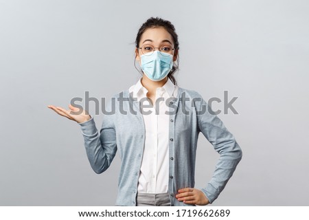 Asian woman in glasses and face mask introduce new company product or banner, pointing, holding promo on hand over left side copy space