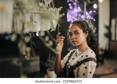 an Asian woman in glasses and wearing overalls standing near a hanging plant in front of a coffee shop terrace at night