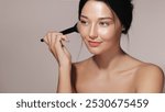 Asian woman with glowing skin applying blush with a makeup brush in a studio setting. Ideal for beauty, cosmetics, and skincare advertisements. High-quality image perfect for promotional and