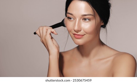 Asian woman with glowing skin applying blush with a makeup brush in a studio setting. Ideal for beauty, cosmetics, and skincare advertisements. High-quality image perfect for promotional and