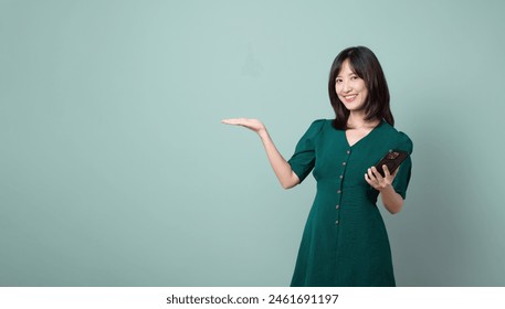 An Asian woman green dress gesturing hand open to free copy space while holding mobile phone isolated on pastel green background. online payment, promotion, deals concept. - Powered by Shutterstock