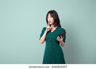 An Asian woman green dress pointing finger shush gesture while holding mobile phone isolated on pastel green background. online payment, promotion, deals concept. - Powered by Shutterstock