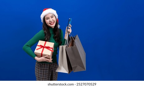 Asian woman in green shirt standing smiling holding phone isolated on blue background, carrying paper bag from Christmas shopping, wearing Santa Claus hat. - Powered by Shutterstock