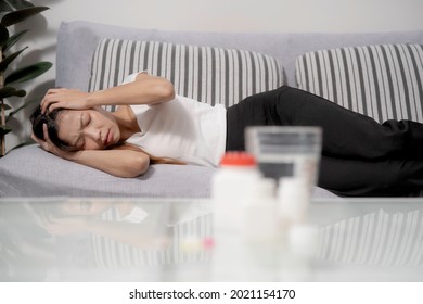 An asian woman had headache, laying down on sofa with blurred medicine and bottle on table, girl put hands on head, fold eye bow together with painful feeling.