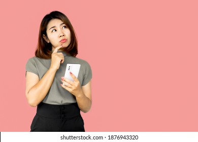 Asian woman hand holding smart phone wonder, thinking and planning something standing over pink background   - Shutterstock ID 1876943320