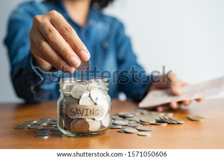 asian woman hand is putting a coin in a glass  bottle and a pile of coins on a brown wooden table,Investment business, retirement, finance and saving money for future concept.