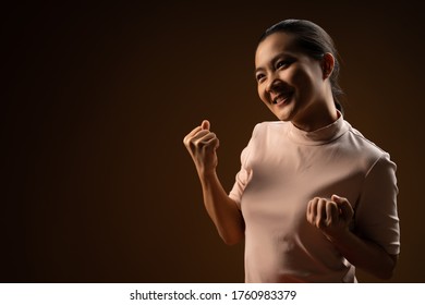 Asian woman happy confident standing showing her fist make a winning gesture isolated on beige background. Low key.