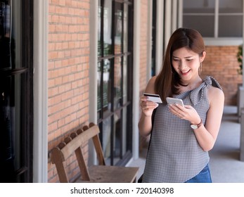 Asian Woman With Happy Face Walking In Shopping Mall While Holding Mobile Phone And Credit Card. Credit Card Use Concept. Online Shopping Concept. Internet Banking Concept