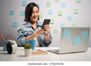 Asian Woman Happy Smiling Looking At Smart Phone And Reading News At Home Office. WFH. Work From Home. Prevention Coronavirus COVID-19 Concept.