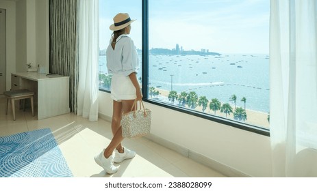 Asian woman with a hat standing by the window of a hotel room with bright fresh colors, a minimal style bedroom with an ocean view, women looking out from the window at the ocean  - Powered by Shutterstock