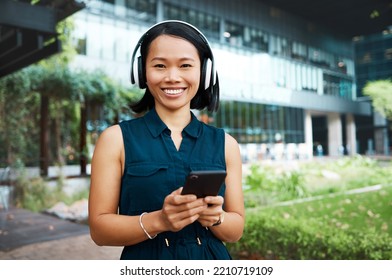 Asian Woman, Headphones And Phone While Streaming Music, Videos Or Podcast Standing Outside On Break Feeling Free And Happy On Internet. Portrait And Smile Of Happy Business Female Using Technology