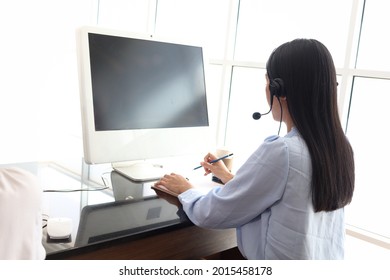 Asian Woman With Headphones Work At Call Center Service Desk Consultant From Behind, Call Center Agent With Headset Talk With Customer On Hotline, Operator With Hands-free Phone Has Conversation