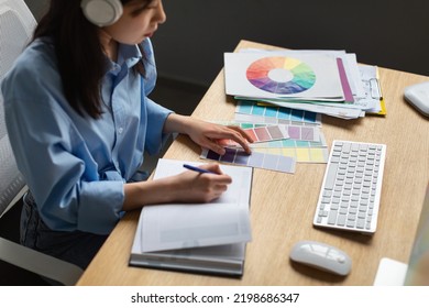 Asian Woman With Headphones Writing Learning Graphic Design Online Working With Color Samples Sitting At Desk Indoor. Designer Working Taking Notes At Workplace In Office. Cropped, Side View