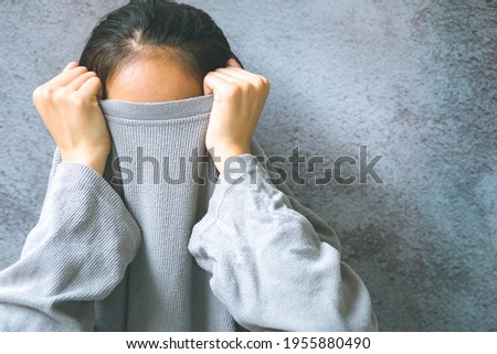 Asian woman hiding face under the clothes. She is pulling a sweater on her head. She has social phobia, trying to be anonymous.