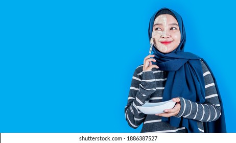 Asian Woman With Hijab On Head And Facial Mask Looking At The Copyspace Over Blue Background. Woman Spa Mask Half-face Beauty Concept.