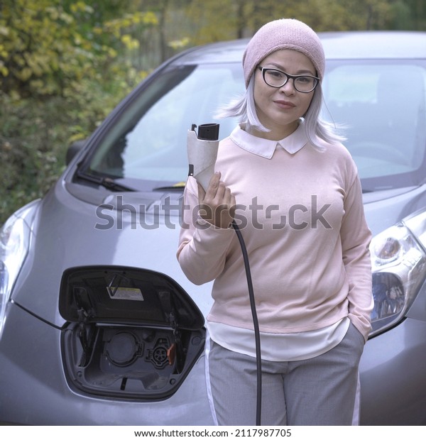 Asian woman holding charging\
cable for electric car at the charge station. Voltage and Power\
concept. Pit stop at charging station. Public EV charger\
station.