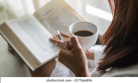 Asian woman holding cup of coffee and reading a book beside the window after get up in morning. Morning lifestyle concept.
