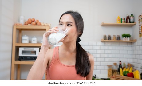 Asian Woman Holding And Drinking Fresh Milk From Glass. Healthy Diet After Exercise In Kitchen, Sport, Health ,exercising, Lifestyle, Healthy Concept.Smiling Thai Female  Happy After Yoga Class.