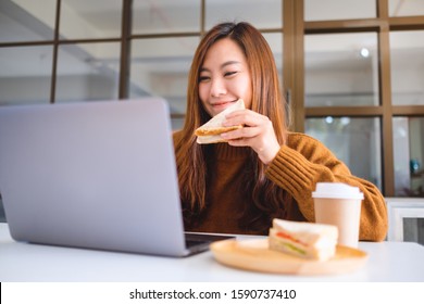 An Asian Woman Holding And Eating Whole Wheat Sandwich While Working On Laptop Computer