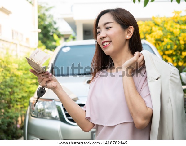 Asian woman holding money and car key\
against a car. insurance, loan and finance\
concept