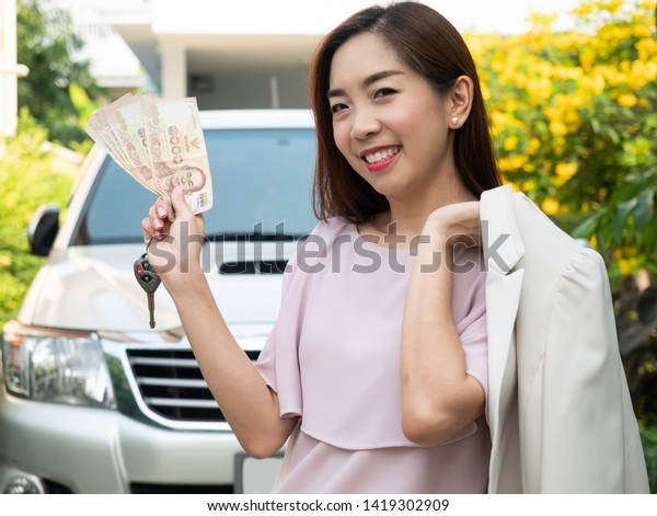 Asian woman holding money and car key
against a car. insurance, loan and finance
concept