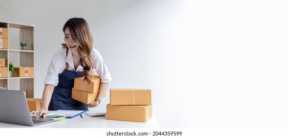 Asian Woman Holding Parcel Boxes And Checking Orders From Laptop, She Owns An Online Store, She Packs And Ships Through A Private Transport Company. Online Selling And Online Shopping Concepts.