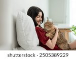 Asian woman holding and play with little cat with happiness at home. Attractive beautiful female lying down on bed, spend leisure time hug and massaging her pet animal with gentle in bedroom in house.