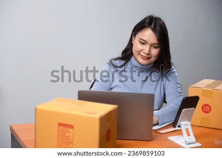 Asian woman holds a parcel box, packs it and ships it through a private courier company. and check orders from laptop She owns an online store.